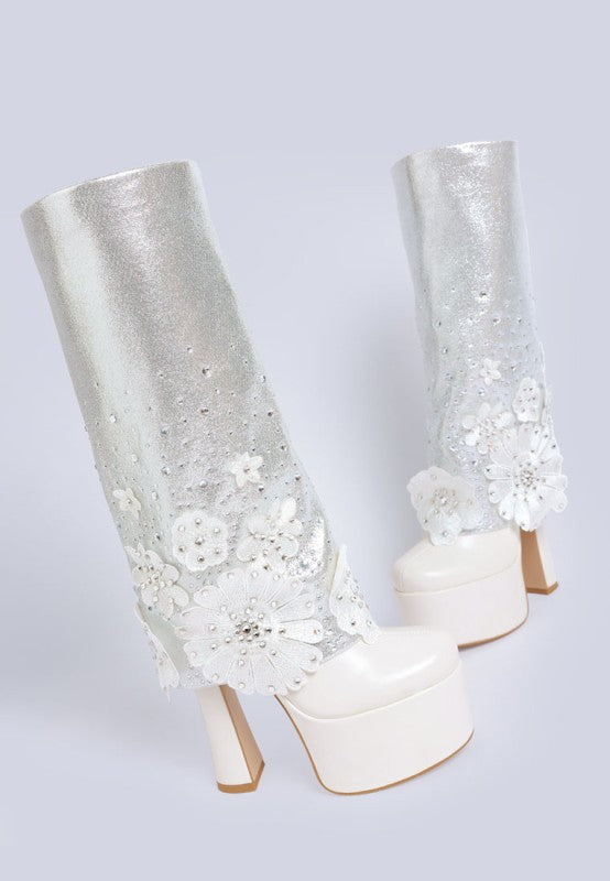A rear three-quarter view of two white 'Sundra' platform boots, showcasing the chunky block heels, the high platforms, and the sparkling glitter shaft adorned with white floral appliques and rhinestones. Available from CO-Wear in Denver, Colorado USA.