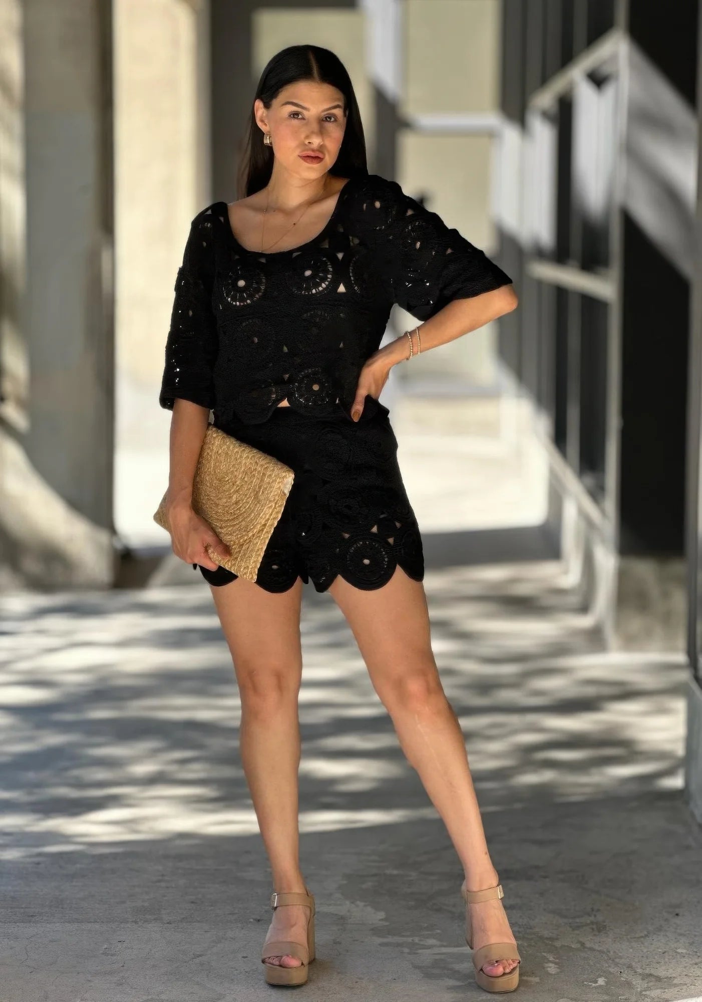 Model wearing a black crochet lace two-piece set, featuring a scoop-neck crop top and matching shorts with scalloped hems, holding a woven clutch. Denver, Colorado USA.