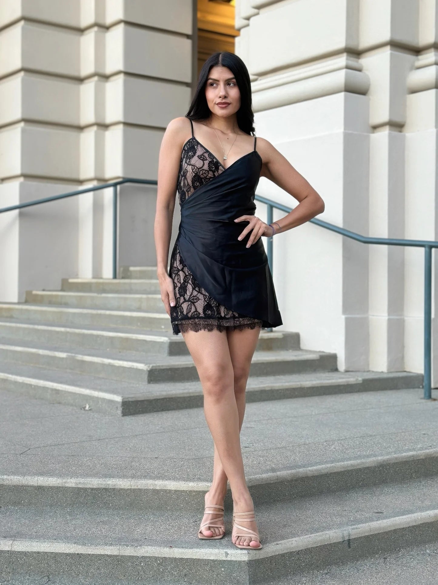Model standing on stone steps in a black mini dress featuring a faux satin wrap overlay over nude lining with black lace detailing on the skirt and bodice. Denver, Colorado USA.