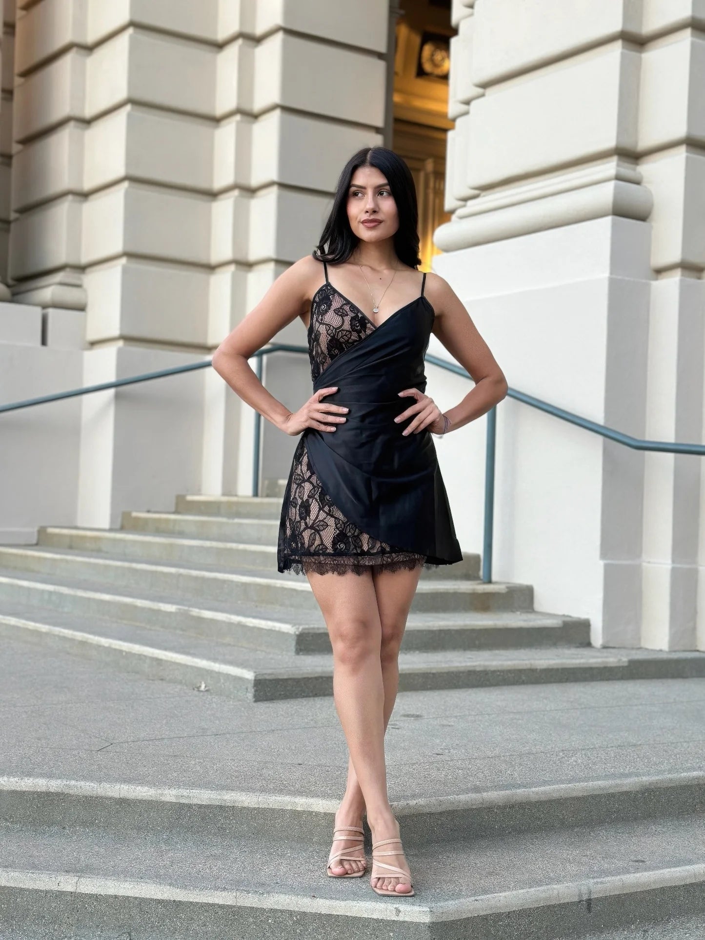 Model standing on stone steps in a black mini dress featuring a satin-like faux wrap overlay over nude lining with sheer black lace paneling on the sides of the bodice and skirt. Denver, Colorado USA.