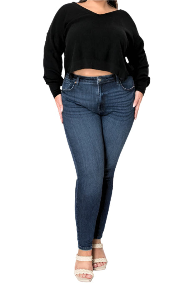 Front view of a plus-size model wearing dark wash super skinny jeans and a black cropped sweater. The jeans have a high-waisted design. The model is also wearing light-colored strappy heels. The focus is on the fit of the jeans, highlighting a sculpted silhouette.