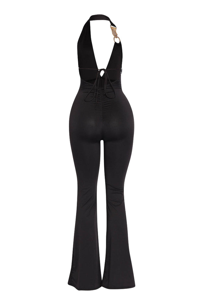
                  
                    A black halter jumpsuit is displayed from the back, featuring a deep V-neckline that extends to the lower back with crisscross tie details. There is a gold-toned ring accent on the right shoulder strap. The jumpsuit has ruched detailing on the lower back and wide flare pants. It is shown on a mannequin or stand against a white background.
                  
                
