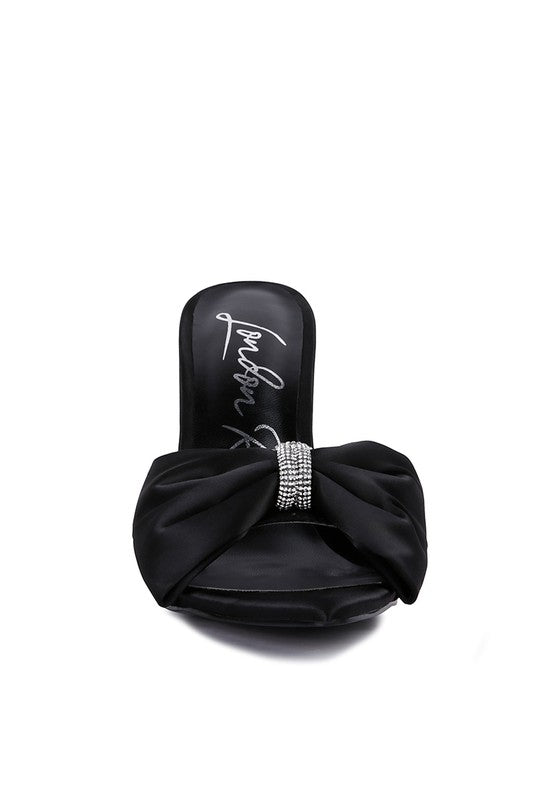 A direct front view of the black satin high-heeled sandal, showcasing the prominent ruched satin bow centered with a sparkling rhinestone band and the pointed open toe design. Available from CO-Wear in Denver, Colorado USA.