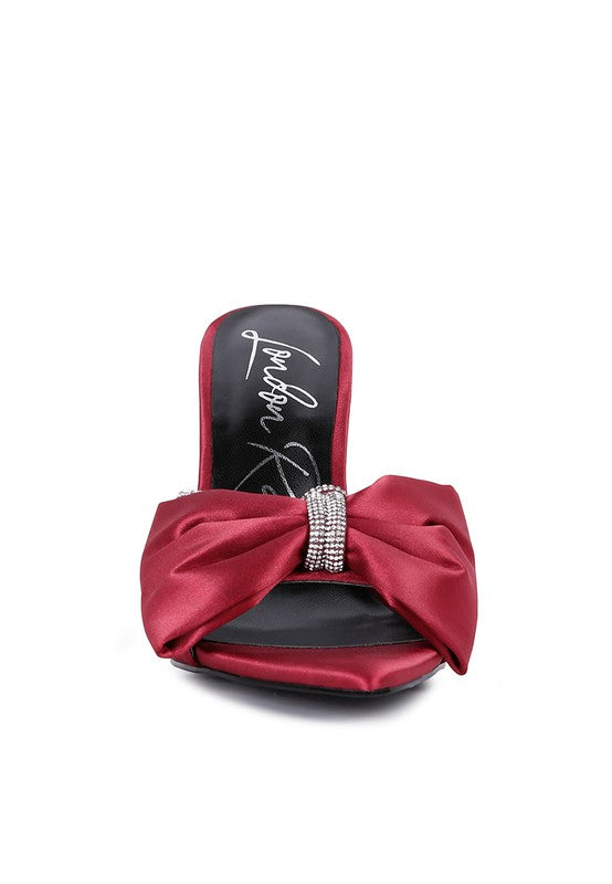 A direct front view of the vibrant red satin high-heeled sandal, showcasing the prominent ruched satin bow centered with a sparkling rhinestone band and the pointed open toe design. Available from CO-Wear in Denver, Colorado USA.