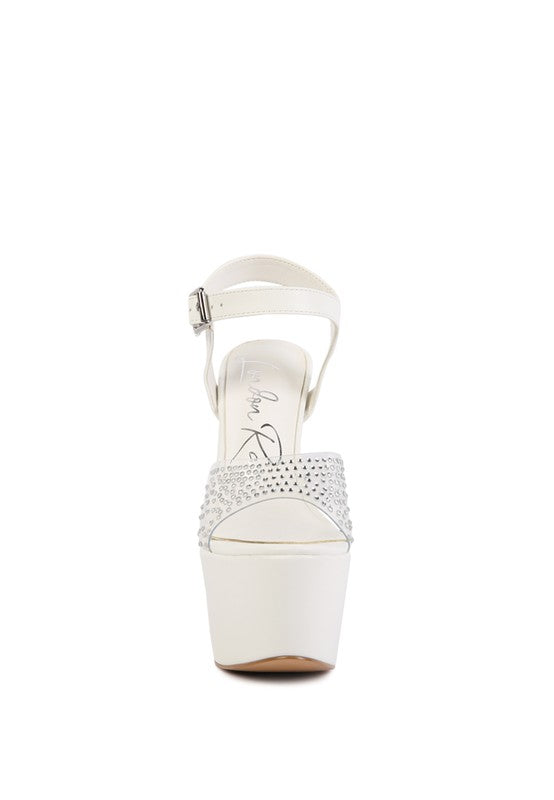 A direct front view of the white 'First Date' sandal, highlighting the massive white platform, the clear toe strap encrusted with rows of rhinestones, and the ankle strap. Available from CO-Wear in Denver, Colorado USA.
