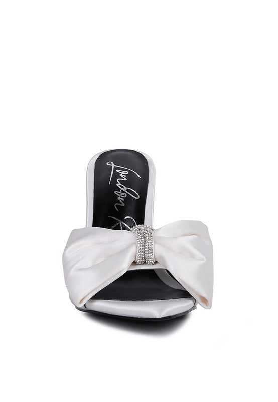 A direct front view of the white satin high-heeled sandal, showcasing the prominent ruched satin bow centered with a sparkling rhinestone band and the pointed open toe design. Available from CO-Wear in Denver, Colorado USA.