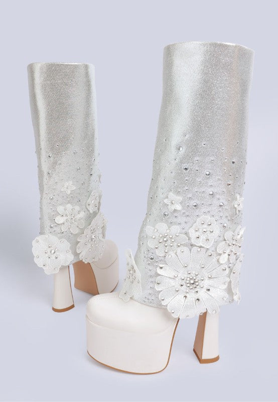 A front view of two white 'Sundra' platform boots, featuring a tall glittery shaft, a thick white platform sole, and large white three-dimensional floral appliques with rhinestone accents. Available from CO-Wear in Denver, Colorado USA.