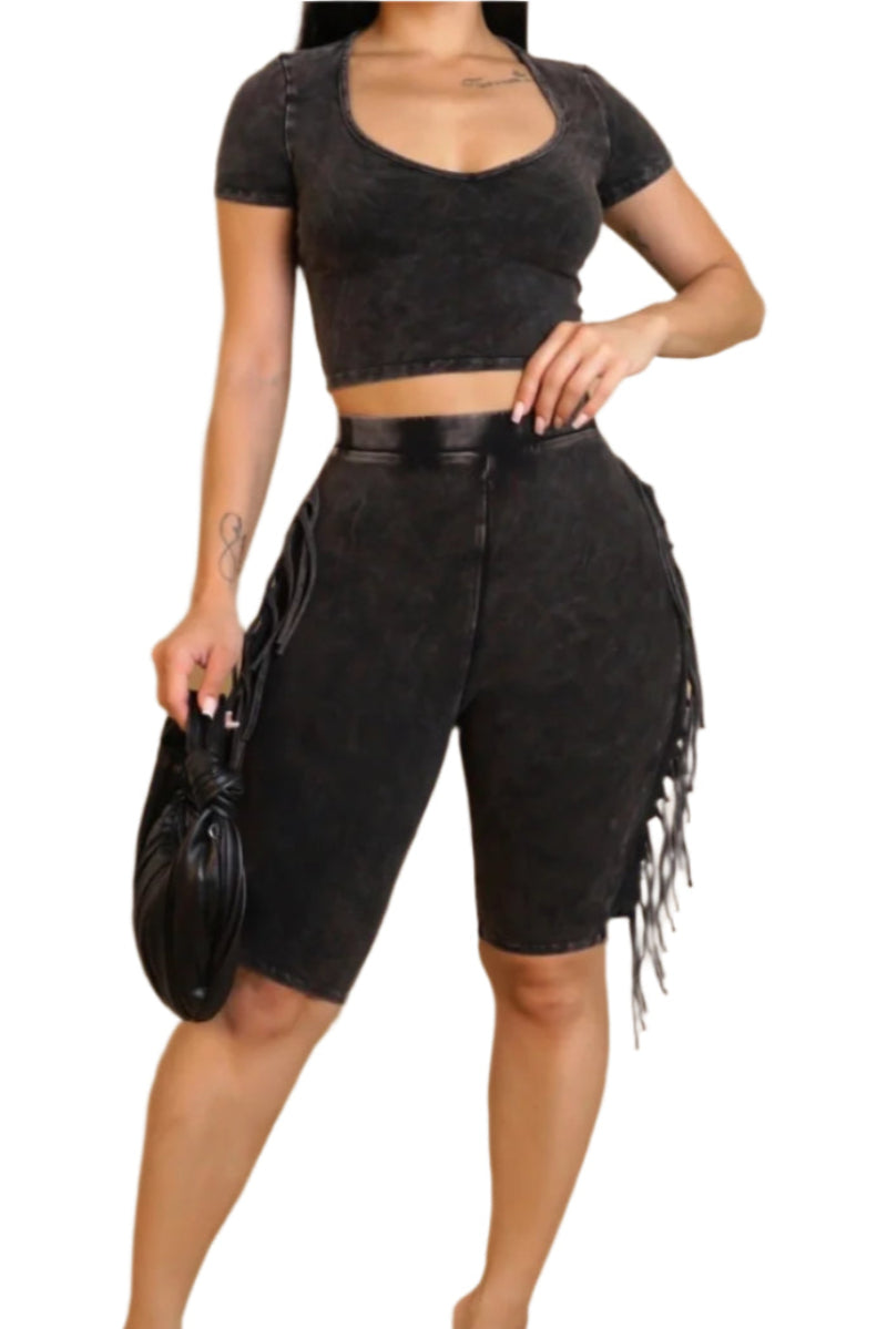 Full body front view of a person wearing a matching black acid-wash two-piece outfit set. The set includes a short-sleeve, scoop-neck crop top and high-waisted biker shorts with fringe detailing on the sides. The person is holding a small black handbag.