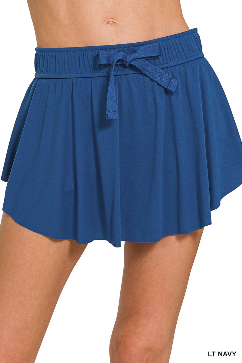A close-up front view of a light navy blue colored skort with a wide elastic waistband and a ruffled, asymmetrical hem with a tie detail on the front. The label "LT NAVY" is visible in the bottom right corner.