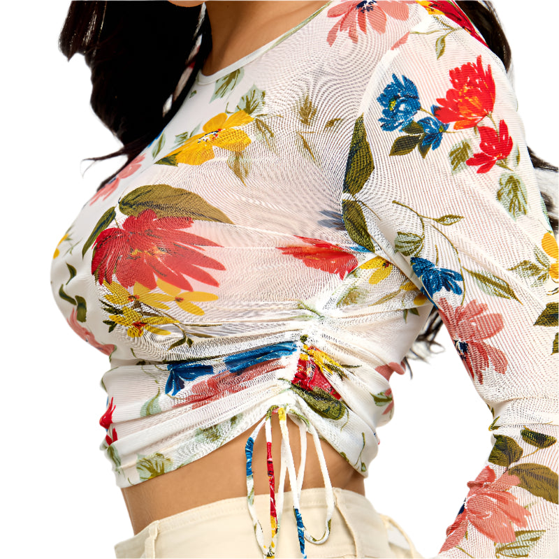 A close-up side view of a white sheer mesh crop top featuring a vibrant floral print with red, yellow, and blue flowers and green leaves. The top has long sleeves with a slight flare and a drawstring detail at the bottom hem creating a ruched effect. White drawstring ties with red, blue, and yellow accents are visible.