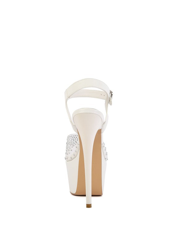 A direct rear view of the white 'First Date' sandal, showcasing the thin stiletto heel, the ankle strap with a buckle, and the clear section of the rhinestone-studded toe strap. Available from CO-Wear in Denver, Colorado USA.