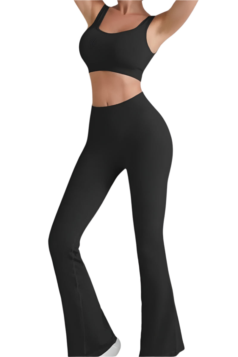 A person is wearing a black, ribbed two-piece activewear set, which includes a sports bra and high-waisted flare leggings. The leggings have a noticeable flare at the bottom and are paired with white sneakers. The background is white.