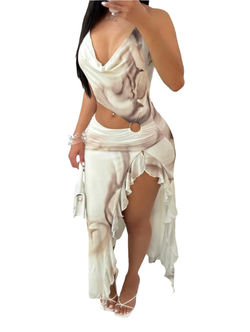 A person is standing, wearing a flowing white and brown abstract print dress with a cowl neck and a high, ruffled slit on the left side. The dress features a cut-out detail at the midriff with a ring accent. The person is holding a white handbag and wearing white strappy heels.