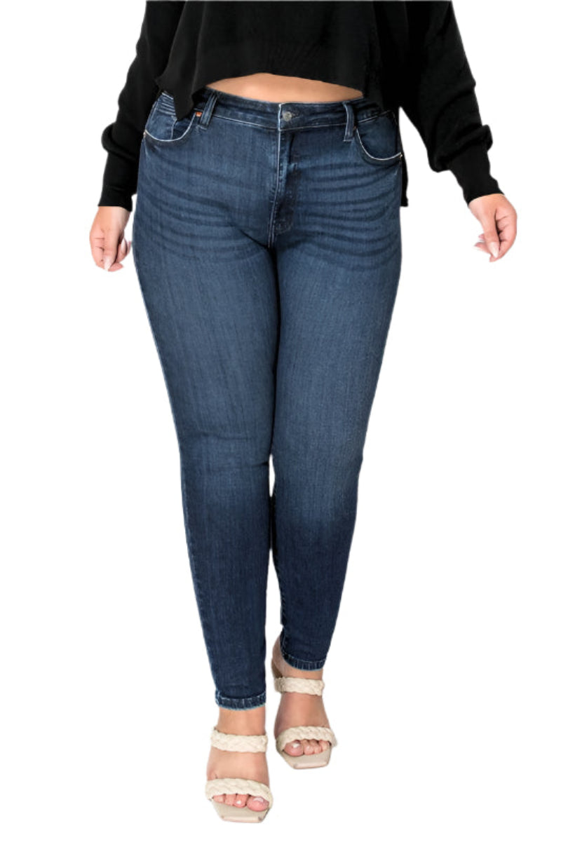 Front view of a plus-size model wearing dark blue super skinny jeans with a high-waisted fit. She is also wearing a black cropped long-sleeve top and light beige woven block-heeled sandals. The jeans are designed to enhance curves, providing a sleek silhouette.
