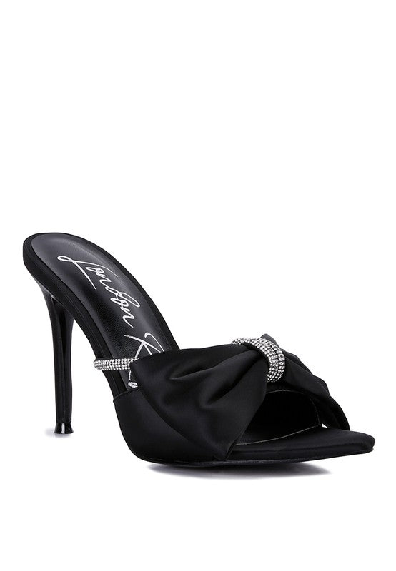 A side profile view of the black satin high-heeled sandal, showcasing the tall, sleek stiletto heel, the ruched satin bow embellished with rhinestones, and the open back mule style. Available from CO-Wear in Denver, Colorado USA.