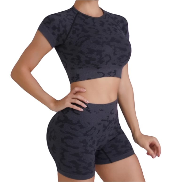 A person is wearing a dark grey or black activewear set with a subtle camouflage pattern. The set includes a short-sleeve crop top and high-waisted booty shorts. The background is white.