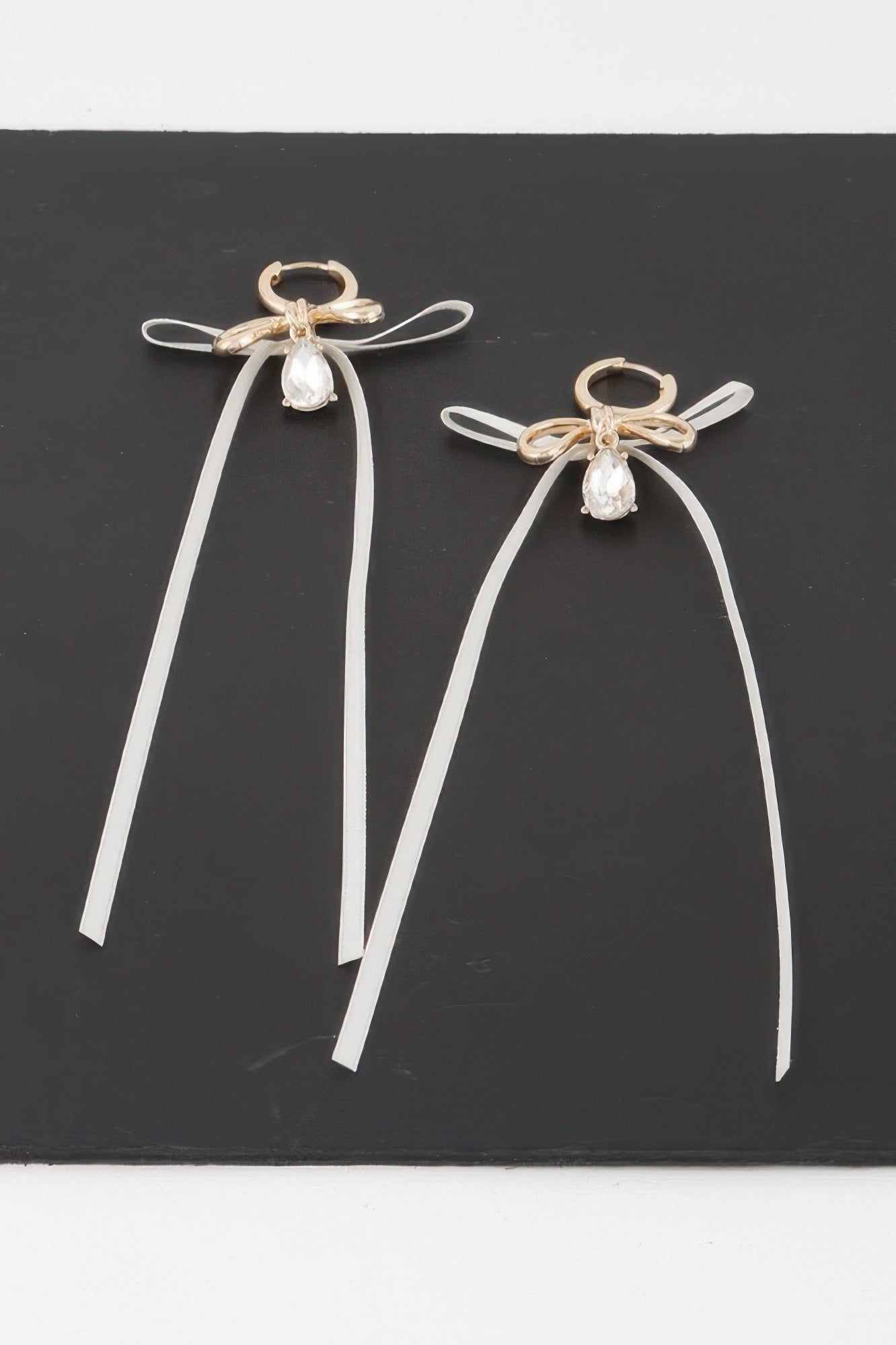 A pair of dangling earrings featuring a gold-toned ribbon-wrapped bow, a clear teardrop crystal, and long white vinyl/leather ribbon drops, set on a black background. Available from CO-Wear in Denver, Colorado USA.