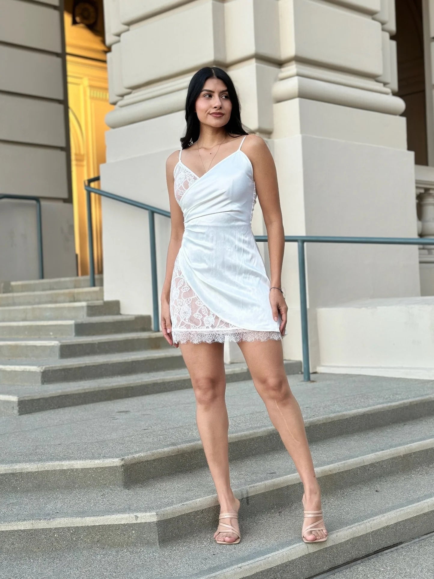 Model standing on stone steps in a white mini dress featuring a satin-like faux wrap over nude lining with sheer white lace detailing on the sides of the skirt. Denver, Colorado USA.