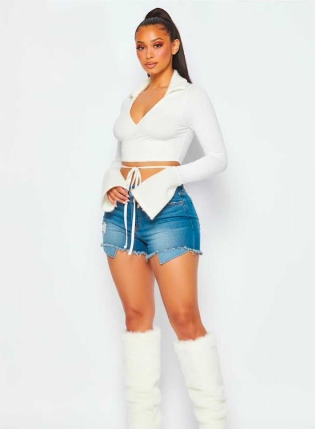 Model wearing a white long-sleeve wrap crop top with flared sleeves, paired with distressed denim shorts and white furry knee-high boots. Denver, Colorado USA.