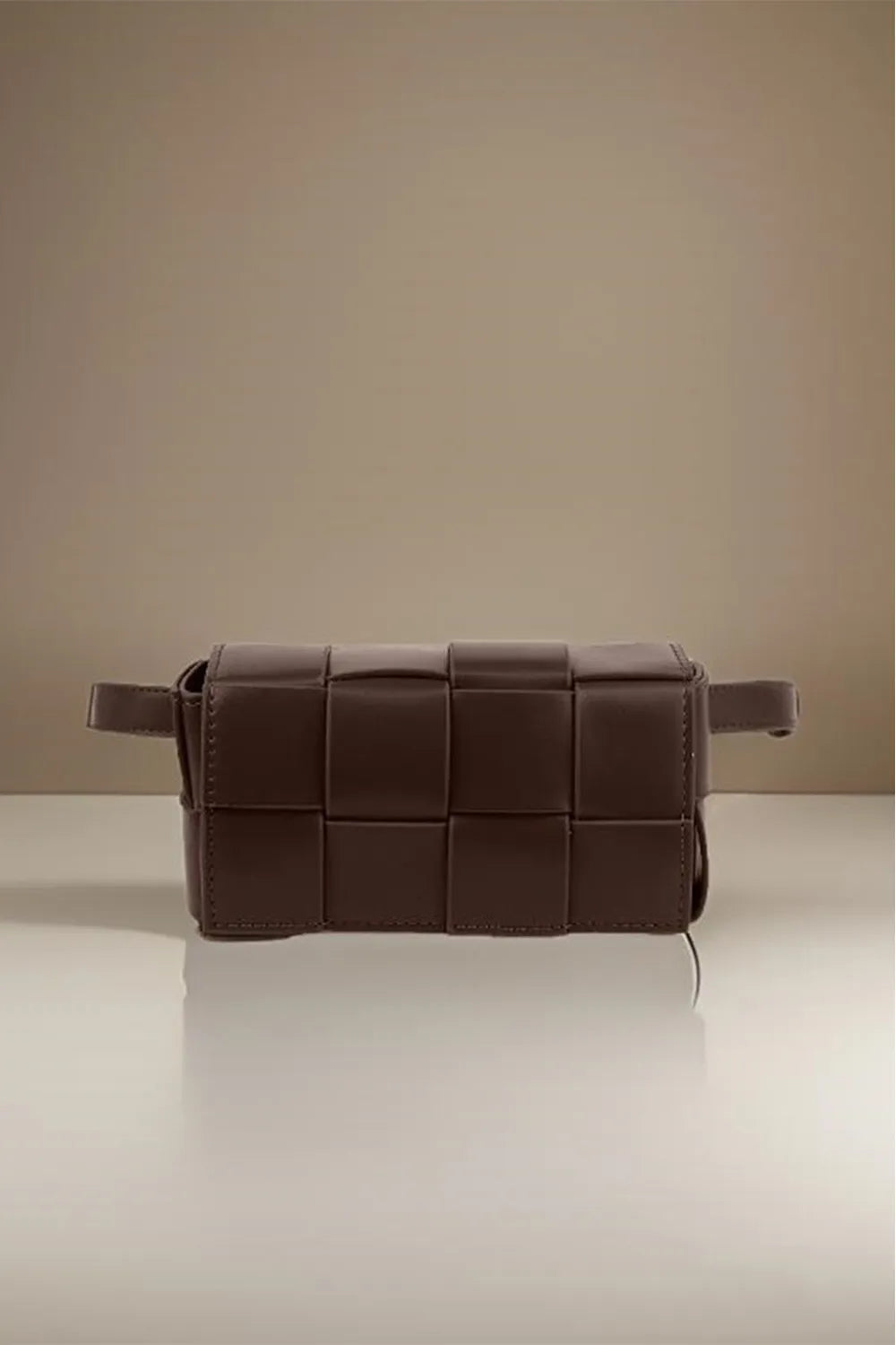 Brown woven leather bag on a reflective surface with a beige background