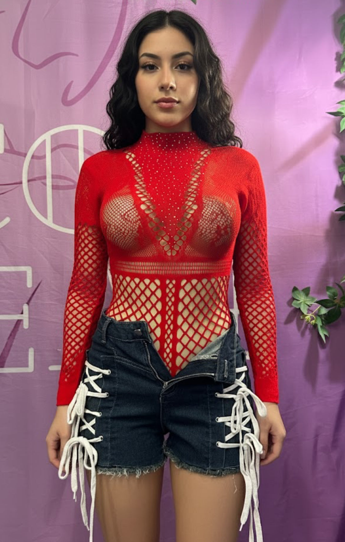 Person wearing a red fishnet top and denim shorts against a purple background